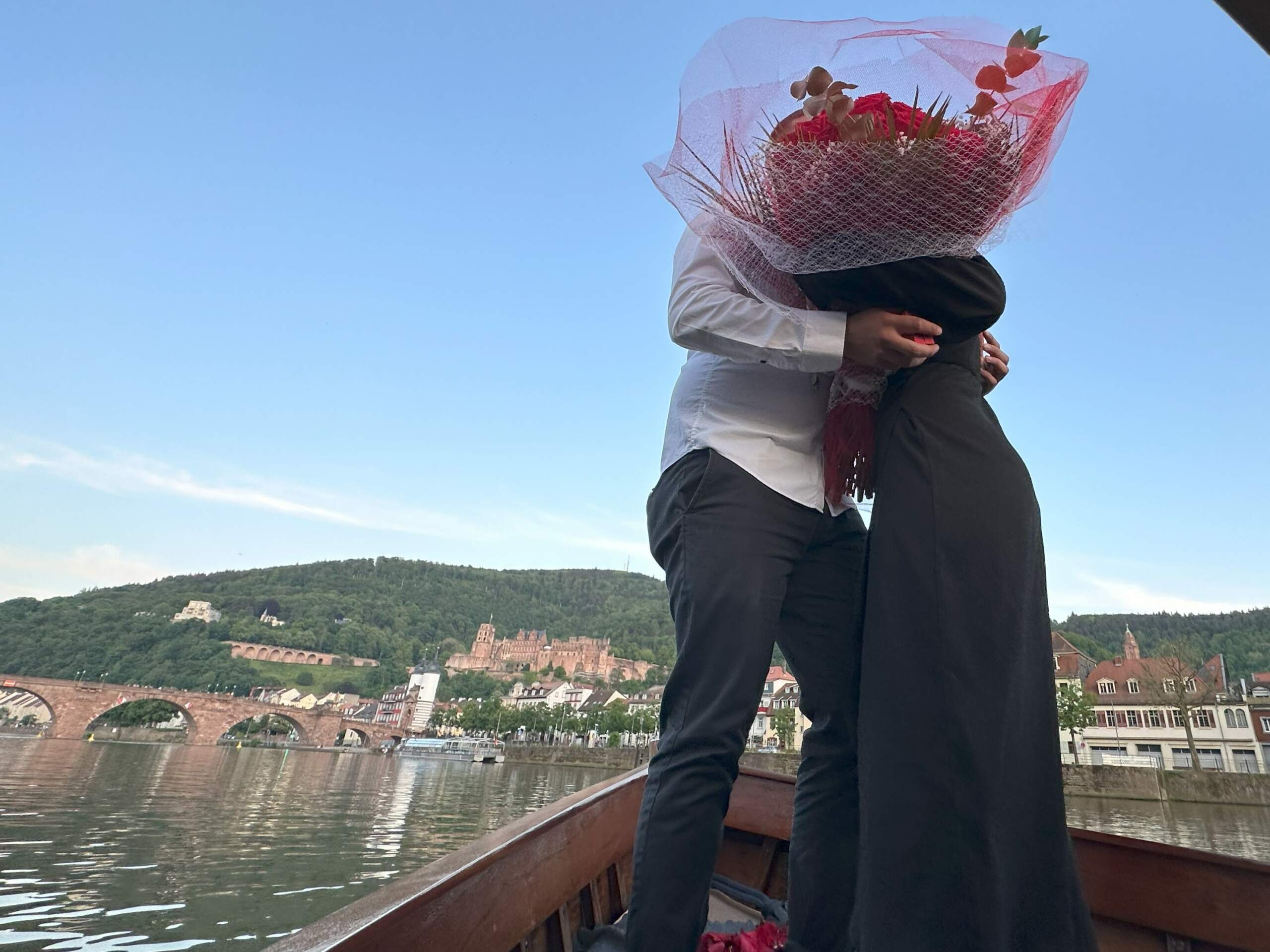 Ein Mann und eine Frauen haben sich an Bord des Bootes von Riverboat verlobt, sie stehen am Bug des Bugs und umarmen sich mit einem Strauß Rosen in der Hand, dahinter das Heidelberger Schloss und die Alte Brücke
